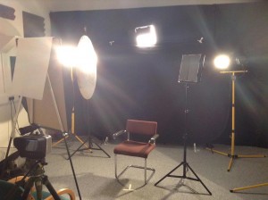 Left to right: the flipchart holding up the key bounce reflector, the halogen key source with the flagging reflector immediately to the right of it, the hair-light LED panel peeking over the backdrop above the hot seat, the LED panel acting as a flag, and the halogen 3/4 backlight.